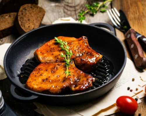 Grilled pork chops in sweet honey glaze, served in grill iron skillet with fresh thyme, rye bread and cherry tomatoes, vertical composition. Food still life. Selective focus